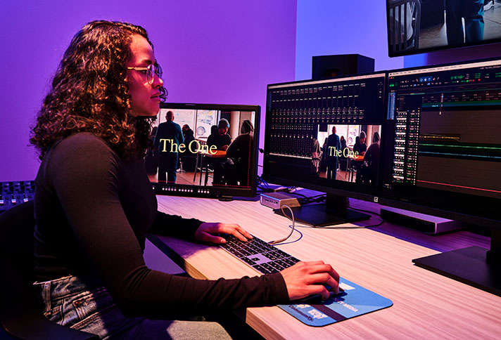 a person with long hair and glasses sitting at a desk with multiple large monitors for editing video