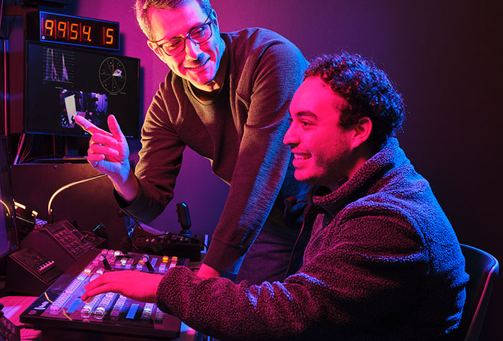 an instructor standing next to a student sitting at a large control panel
