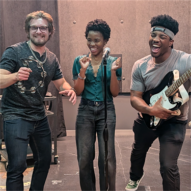 Three performers laugh and pose in a Denver Community Media studio. One holds a tambourine, another stands at a mic, and the third plays electric guitar. All are mid-laugh and vibing.