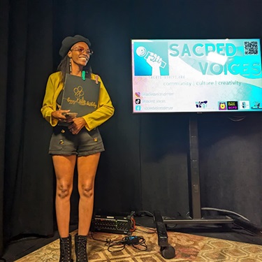 Person standing in DCM’s Main Studio during Sacred Voices event, holding a folder. A screen behind them displays Sacred Voices branding.