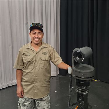 A man named Frankie stands in the Denver Community Media Main Studio next to a PTZ camera, smiling with his hand resting on the camera base.